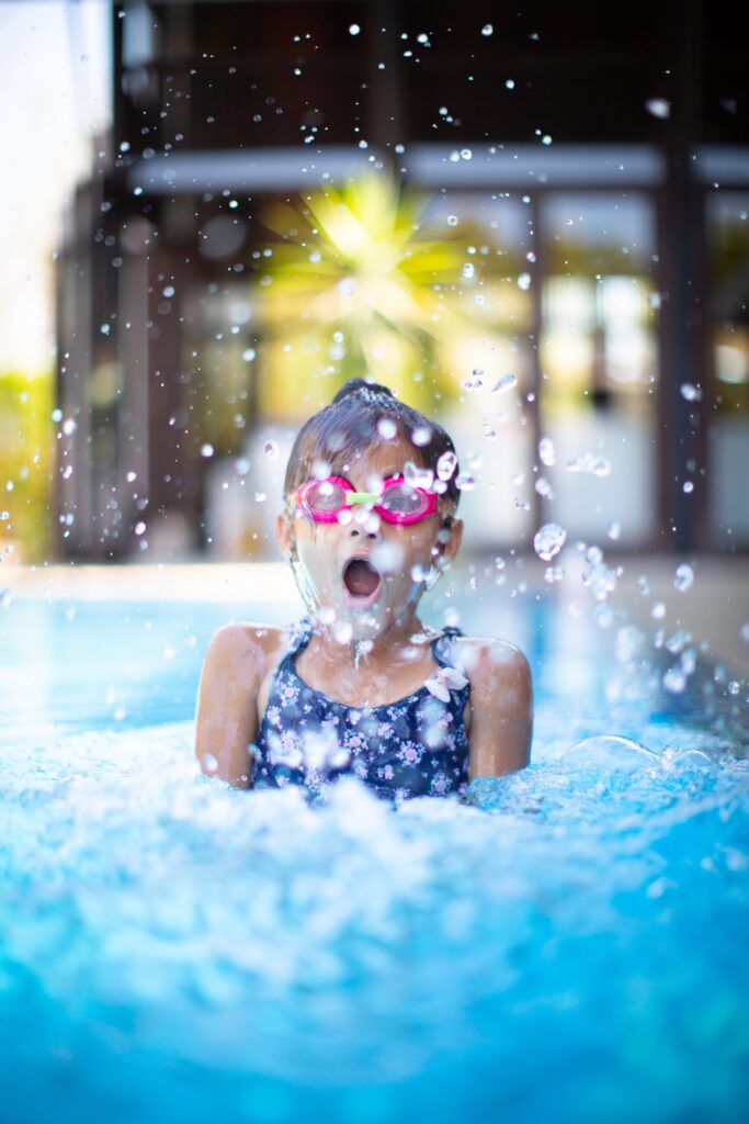 Menina com cerca de 6 anos, usando óculos de natação infantil rosa, mergulhando numa piscina e espelhando água.